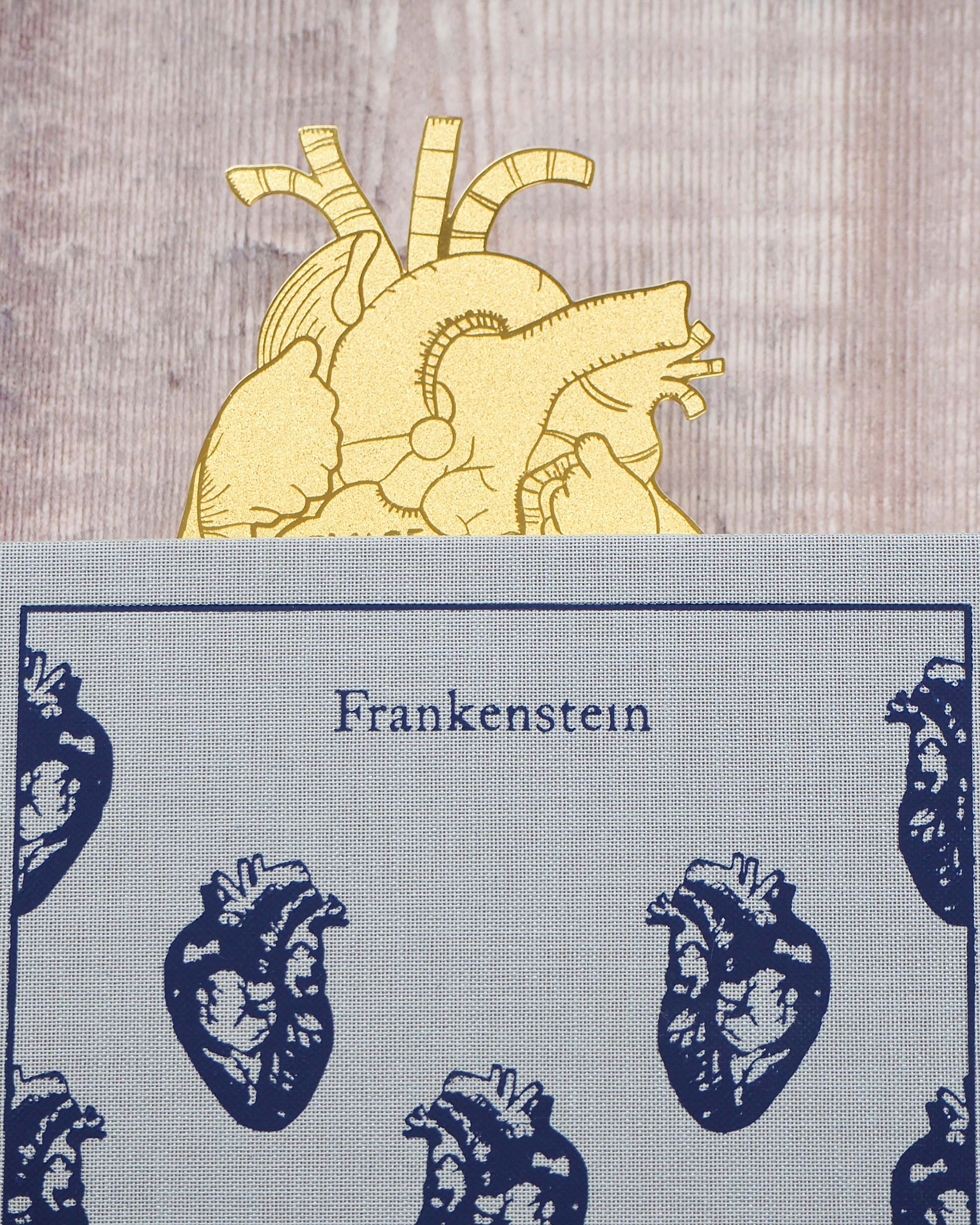 A gold brass anatomical heart-shaped bookmark engraved with the quote “Beware; for I am fearless and therefore powerful” from Mary Shelley’s Frankenstein. The bookmark is delicately etched with intricate vein details, resting on an open book to mark a page.