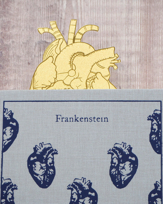 A gold brass anatomical heart-shaped bookmark engraved with the quote “Beware; for I am fearless and therefore powerful” from Mary Shelley’s Frankenstein. The bookmark is delicately etched with intricate vein details, resting on an open book to mark a page.