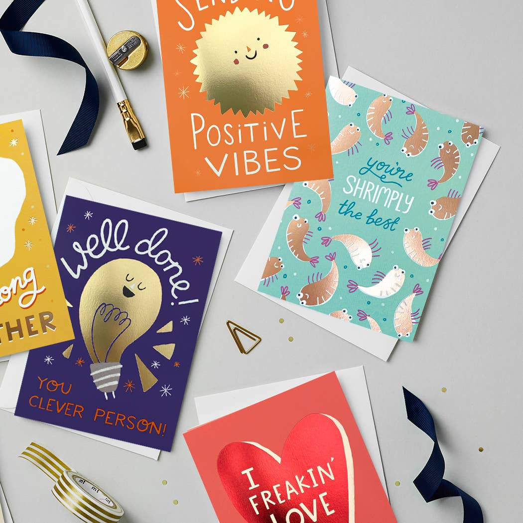 A collection of vibrant and cheerful greeting cards, including a red foil “I Freakin’ Love You” card with a bold heart design. Other cards feature gold foil designs with messages like “Well Done,” “Sending Positive Vibes,” and “You’re Shrimply the Best,” displayed on a flat lay with gold and navy accessories.