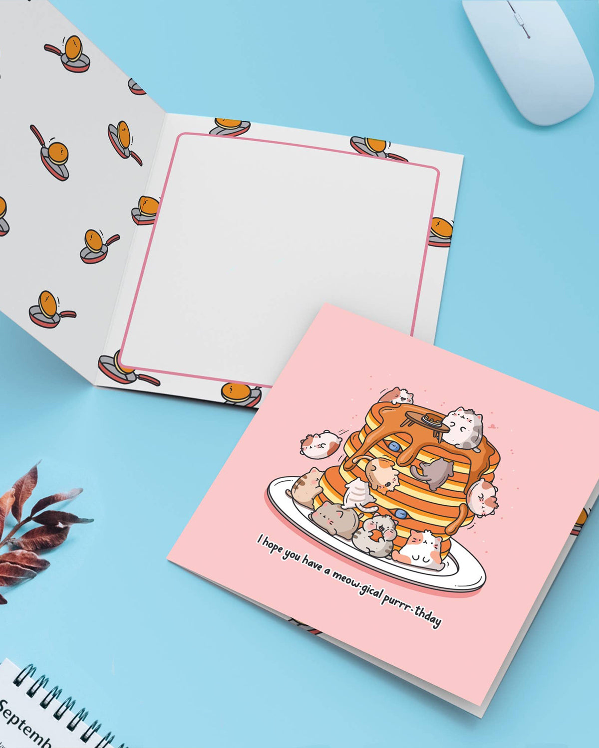 A greeting card featuring cats stacked on pancakes sits open to reveal its patterned interior and message space. Surrounding are cute props on a pastel background. A sweet and punny Toastedink card stocked by Rebel Romance for magical birthdays.