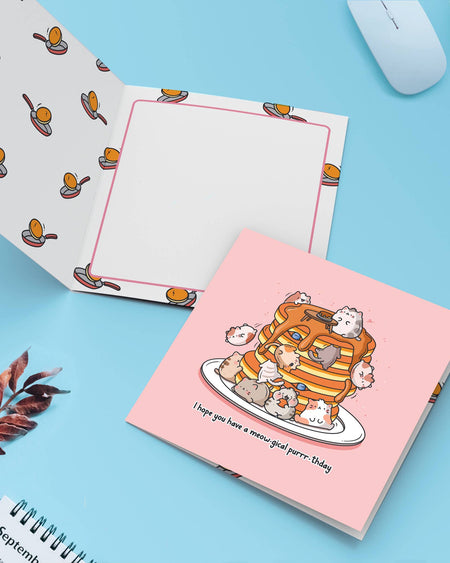A greeting card featuring cats stacked on pancakes sits open to reveal its patterned interior and message space. Surrounding are cute props on a pastel background. A sweet and punny Toastedink card stocked by Rebel Romance for magical birthdays.