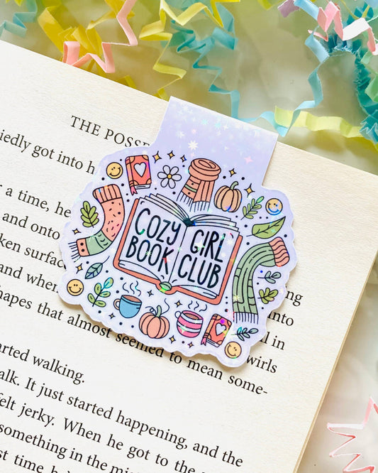 A Rebel Romance “Cozy Girl Book Club” magnetic bookmark by The Stickityboo Co holding a page in an open book. Surrounded by pastel confetti paper, the illustrated design features autumn icons like scarves, mugs and pumpkins, ideal for book lovers.