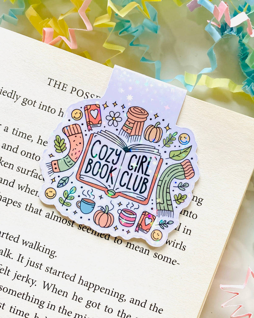 A Rebel Romance “Cozy Girl Book Club” magnetic bookmark by The Stickityboo Co holding a page in an open book. Surrounded by pastel confetti paper, the illustrated design features autumn icons like scarves, mugs and pumpkins, ideal for book lovers.