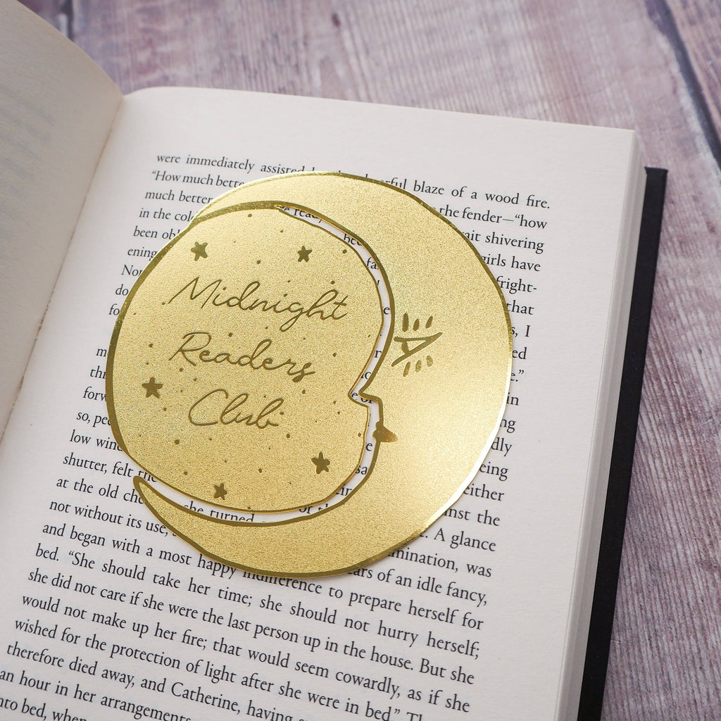 A crescent moon-shaped brass bookmark with “Midnight Readers Club” and celestial stars etched onto its surface, placed on an open book to mark a page. The bookmark adds an elegant and whimsical touch, ideal for book lovers who enjoy night-time reading.