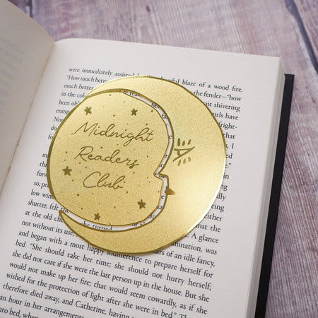 A crescent moon-shaped brass bookmark with “Midnight Readers Club” and celestial stars etched onto its surface, placed on an open book to mark a page. The bookmark adds an elegant and whimsical touch, ideal for book lovers who enjoy night-time reading.