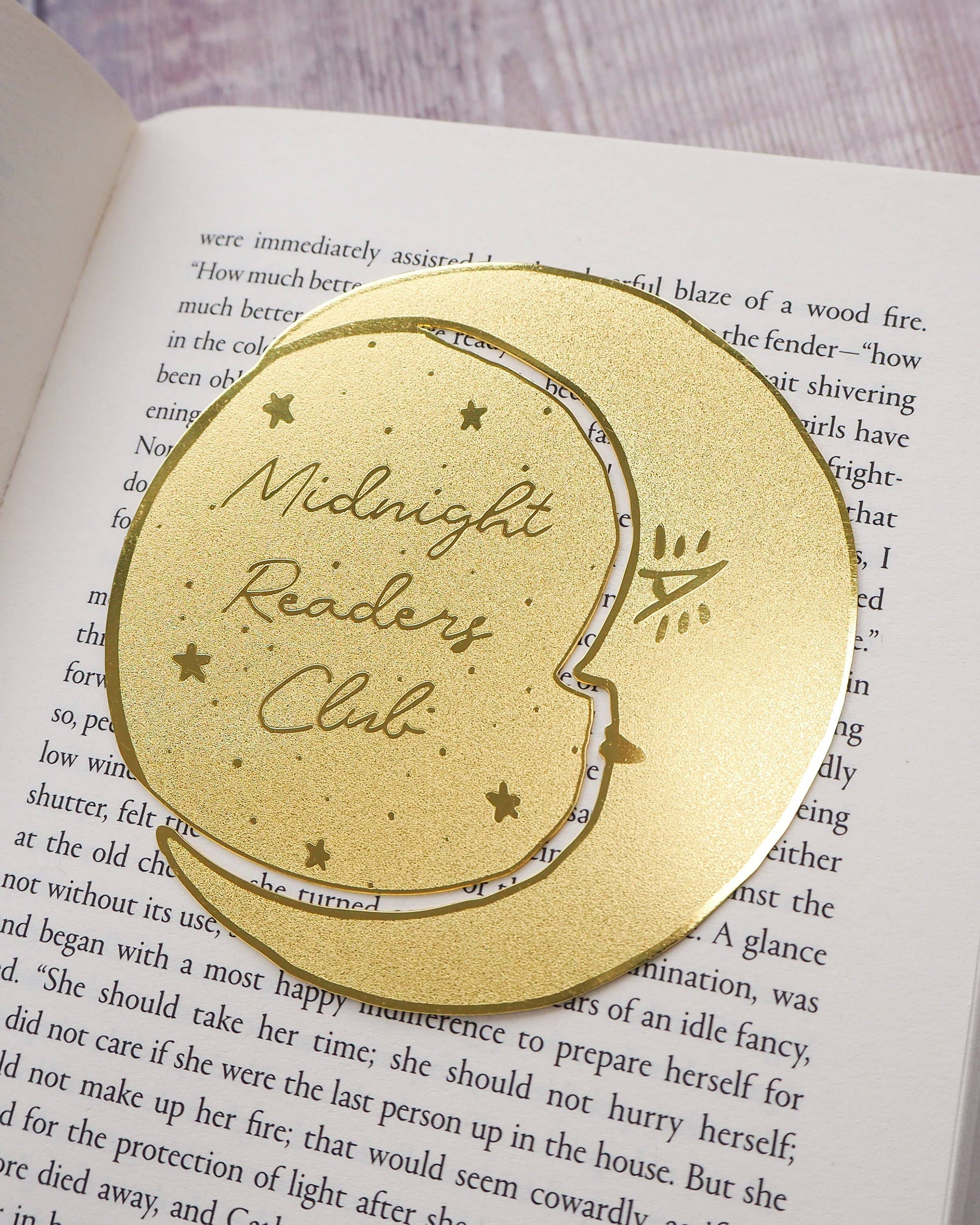 A crescent moon-shaped brass bookmark with “Midnight Readers Club” and celestial stars etched onto its surface, placed on an open book to mark a page. The bookmark adds an elegant and whimsical touch, ideal for book lovers who enjoy night-time reading.