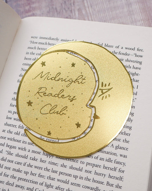 A crescent moon-shaped brass bookmark with “Midnight Readers Club” and celestial stars etched onto its surface, placed on an open book to mark a page. The bookmark adds an elegant and whimsical touch, ideal for book lovers who enjoy night-time reading.