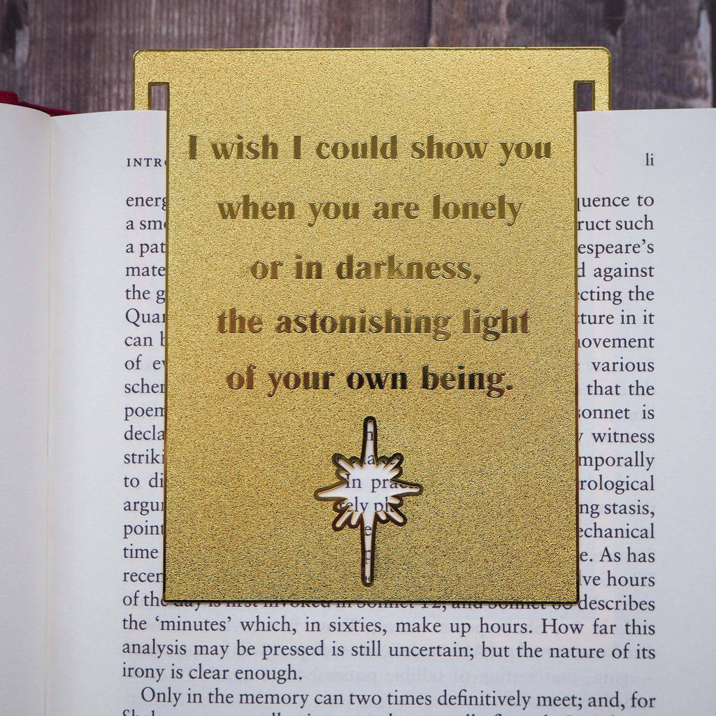 A close-up image of a brass bookmark engraved with the heartfelt Hafiz of Shiraz quote: “I wish I could show you, when you are lonely or in darkness, the astonishing light of your own being.” The bookmark is resting on the pages of an open book, showcasing its intricate detailing and elegant design.