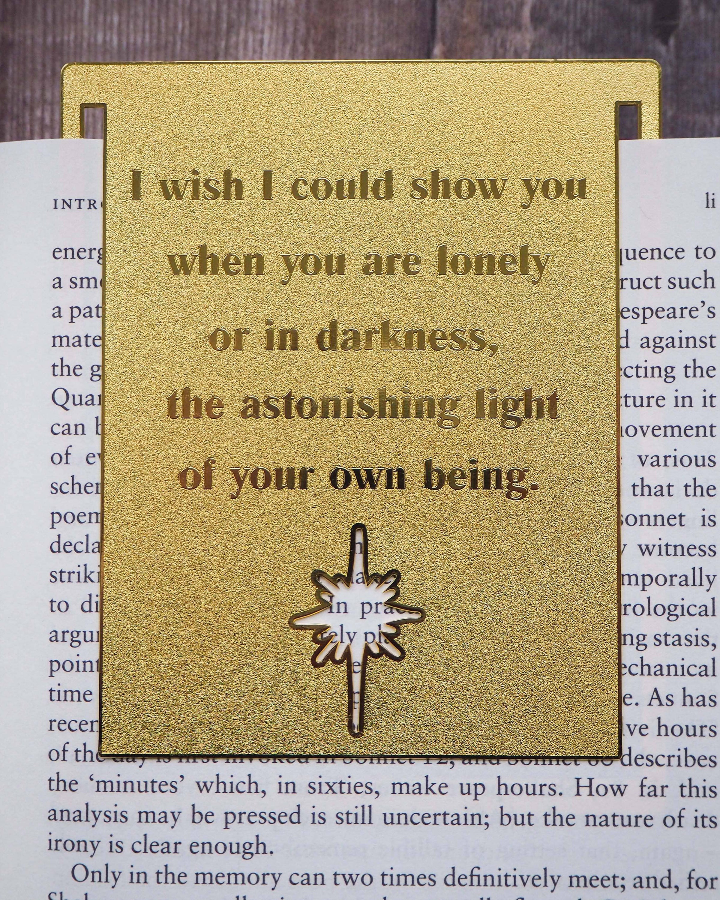 A close-up image of a brass bookmark engraved with the heartfelt Hafiz of Shiraz quote: “I wish I could show you, when you are lonely or in darkness, the astonishing light of your own being.” The bookmark is resting on the pages of an open book, showcasing its intricate detailing and elegant design.