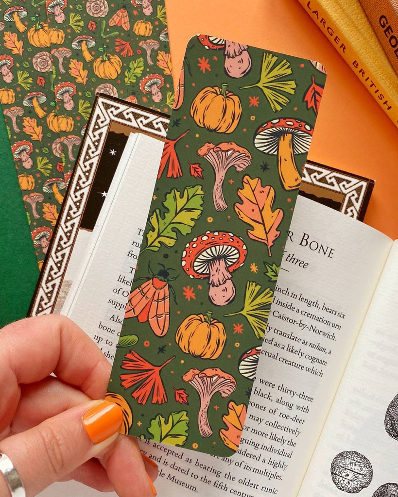 A hand holding an autumnal themed green bookmark filled with hand illustrated autumnal leaves, mushrooms, moths, pumpkins and sparkles. 