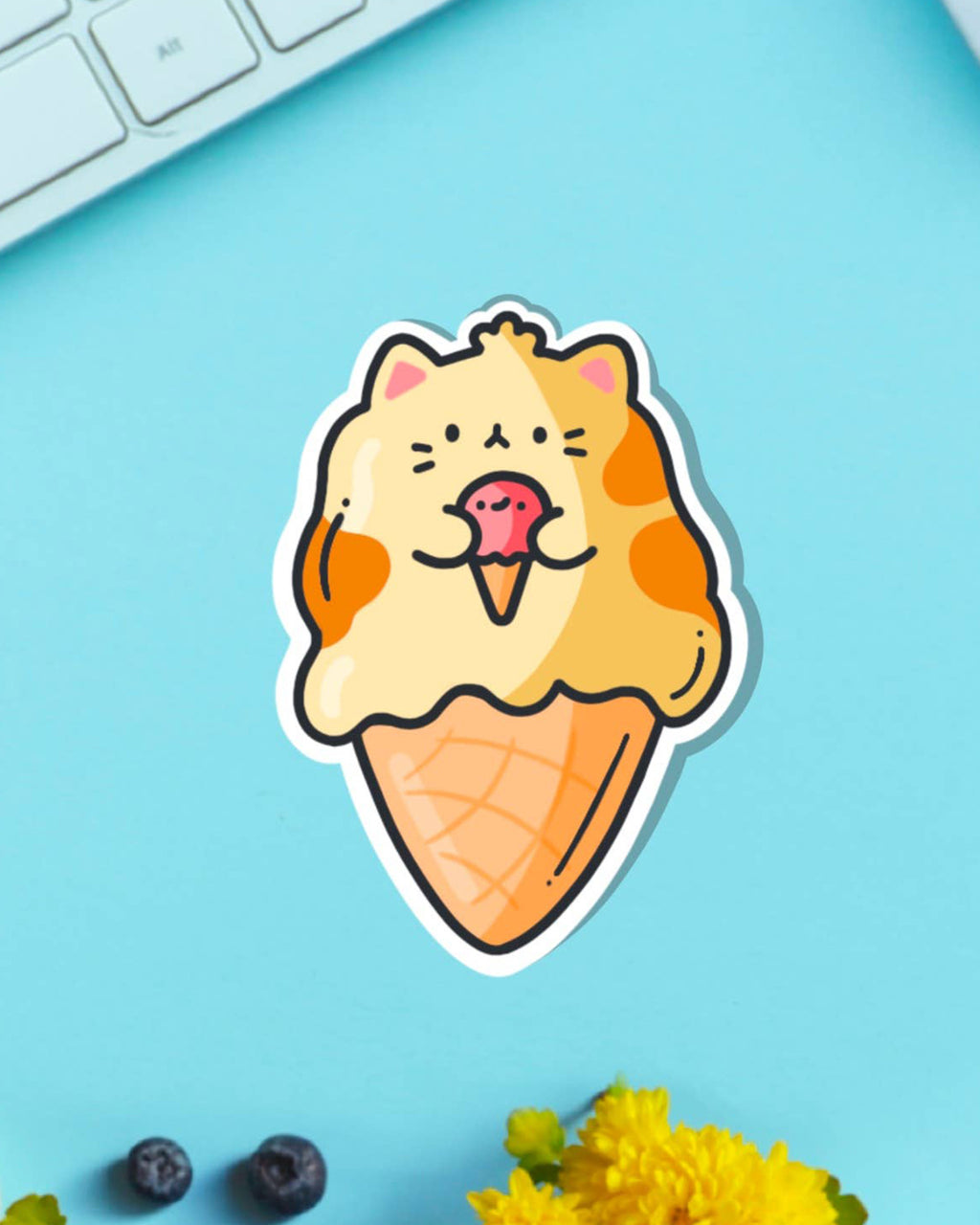 Adorable Toastedink vinyl sticker of a cartoon cat turned into an ice cream cone, placed on a blue desk surface with yellow flowers, blueberries, and a white keyboard nearby.