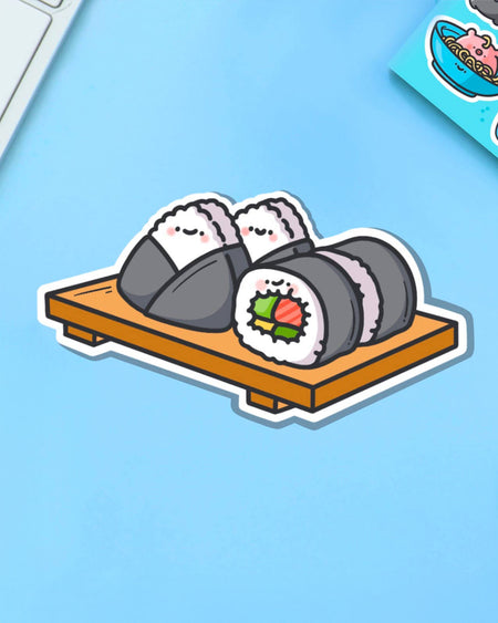 Playful sushi platter vinyl sticker with kawaii-style smiling sushi and onigiri on a tray. Shot on a pale blue background with part of a sticker sheet visible. From Rebel Romance, a UK small biz curating cute, quirky and alternative goods.