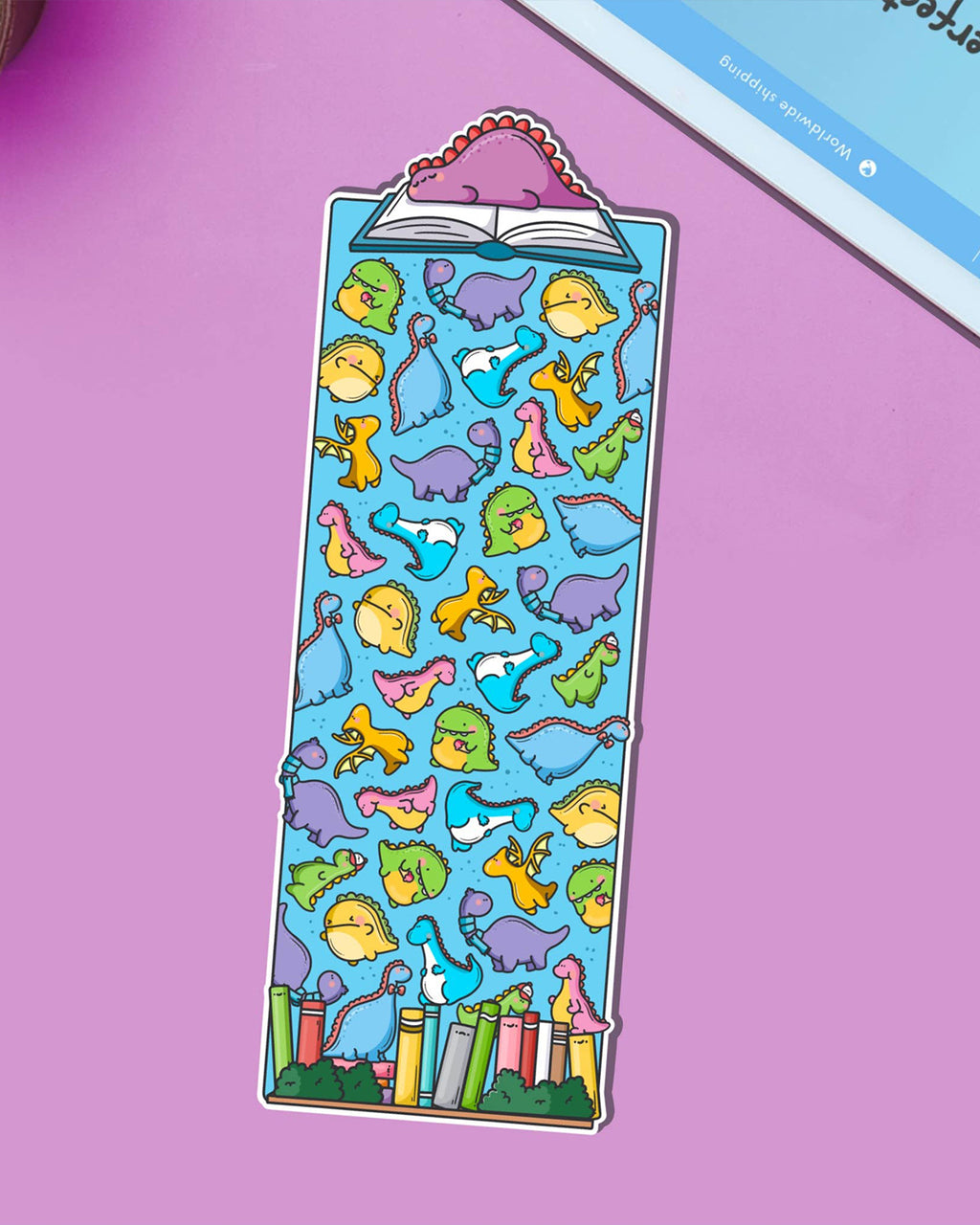 Cute dinosaur bookmark by Toastedink, sold by Rebel Romance, featuring colourful cartoon dinosaurs scattered across a blue background, with a pink dinosaur reading a book at the top. Displayed on a purple surface.