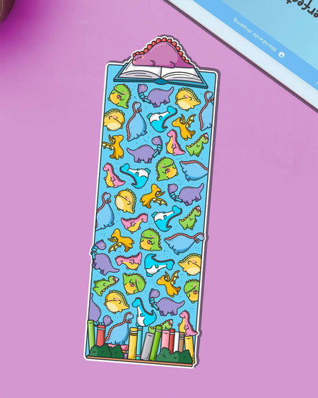 Cute dinosaur bookmark by Toastedink, sold by Rebel Romance, featuring colourful cartoon dinosaurs scattered across a blue background, with a pink dinosaur reading a book at the top. Displayed on a purple surface.