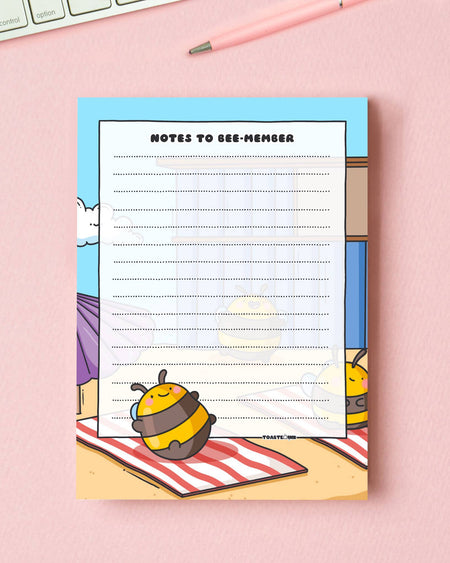 Adorable A6 bee-themed notepad designed by Toastedink, stocked by Rebel Romance. Titled "Notes to Bee-member", with a smiley bumblebee on a picnic blanket. Photographed with a pink pen and keyboard on a pink backdrop.