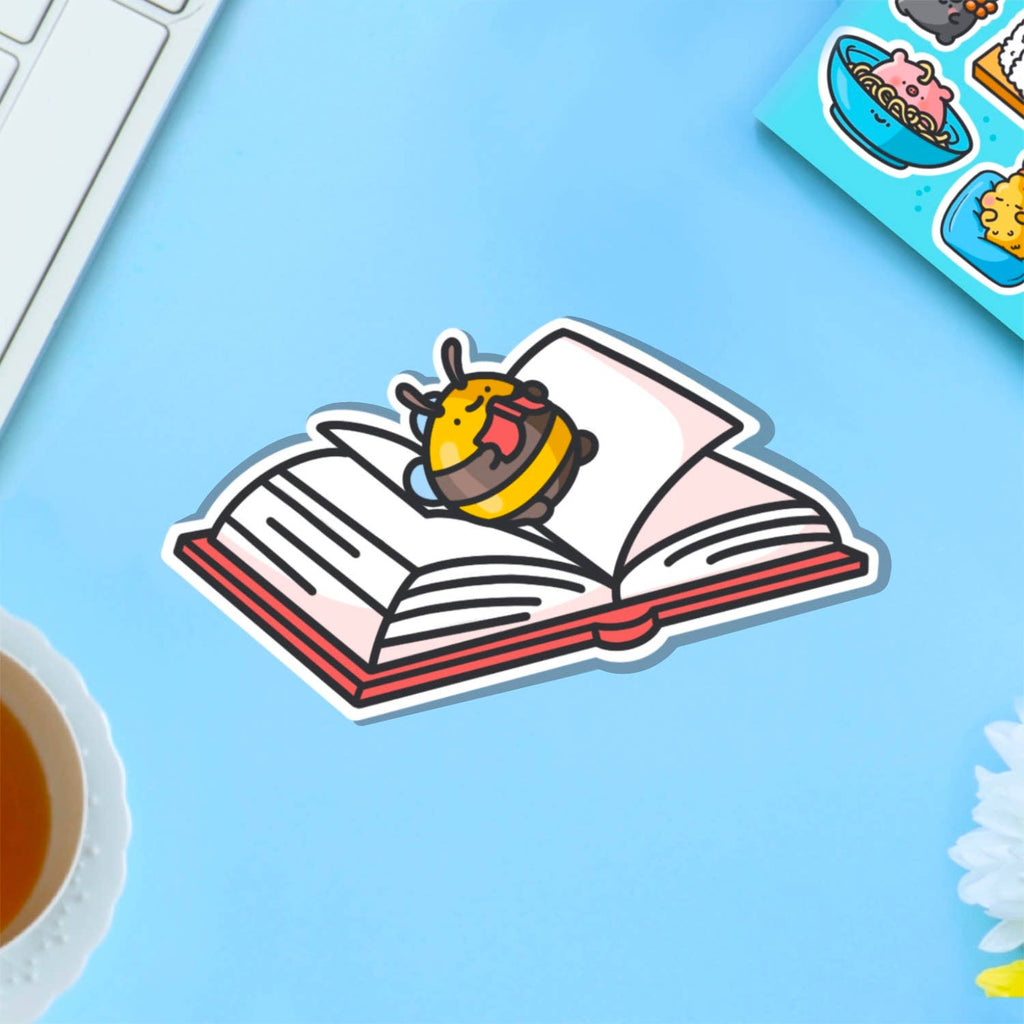 A cute vinyl sticker of a yellow and brown bumblebee lying on its back on top of an open book, tongue sticking out playfully. Displayed on a purple background with pink and blue elements. Glossy waterproof finish, ideal for book lovers and readers.