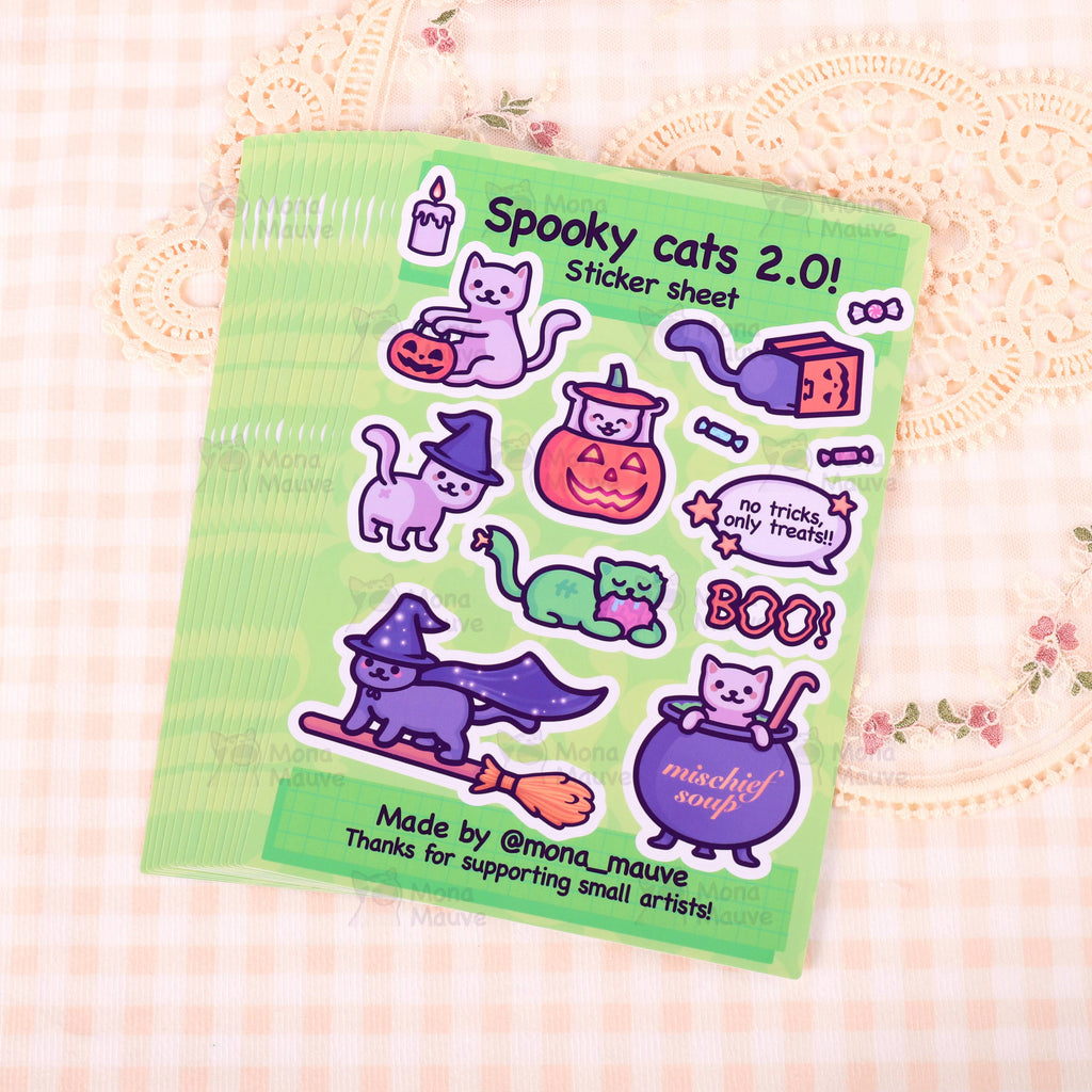 Stack of Spooky Cats 2.0 Sticker Sheets by Mona Mauve laid out on soft patterned fabric. Each sheet includes adorable vinyl Halloween-themed cat illustrations with playful designs like “mischief soup” and cats in pumpkins. A charming spooky-season sticker sheet from a small indie artist.