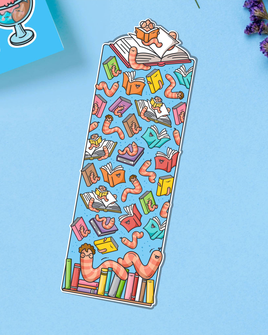 Funny bookworm bookmark by Toastedink at Rebel Romance, with playful pink worms and open books on a sky-blue backdrop. Photo includes sprigs of purple flowers.