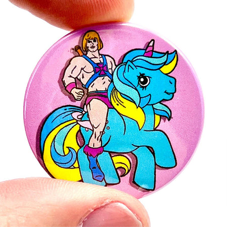 A 38mm high-gloss pin badge featuring He-Man riding a colourful My Little Pony-style unicorn on a pink background. The design combines retro pop culture with a playful, campy vibe.