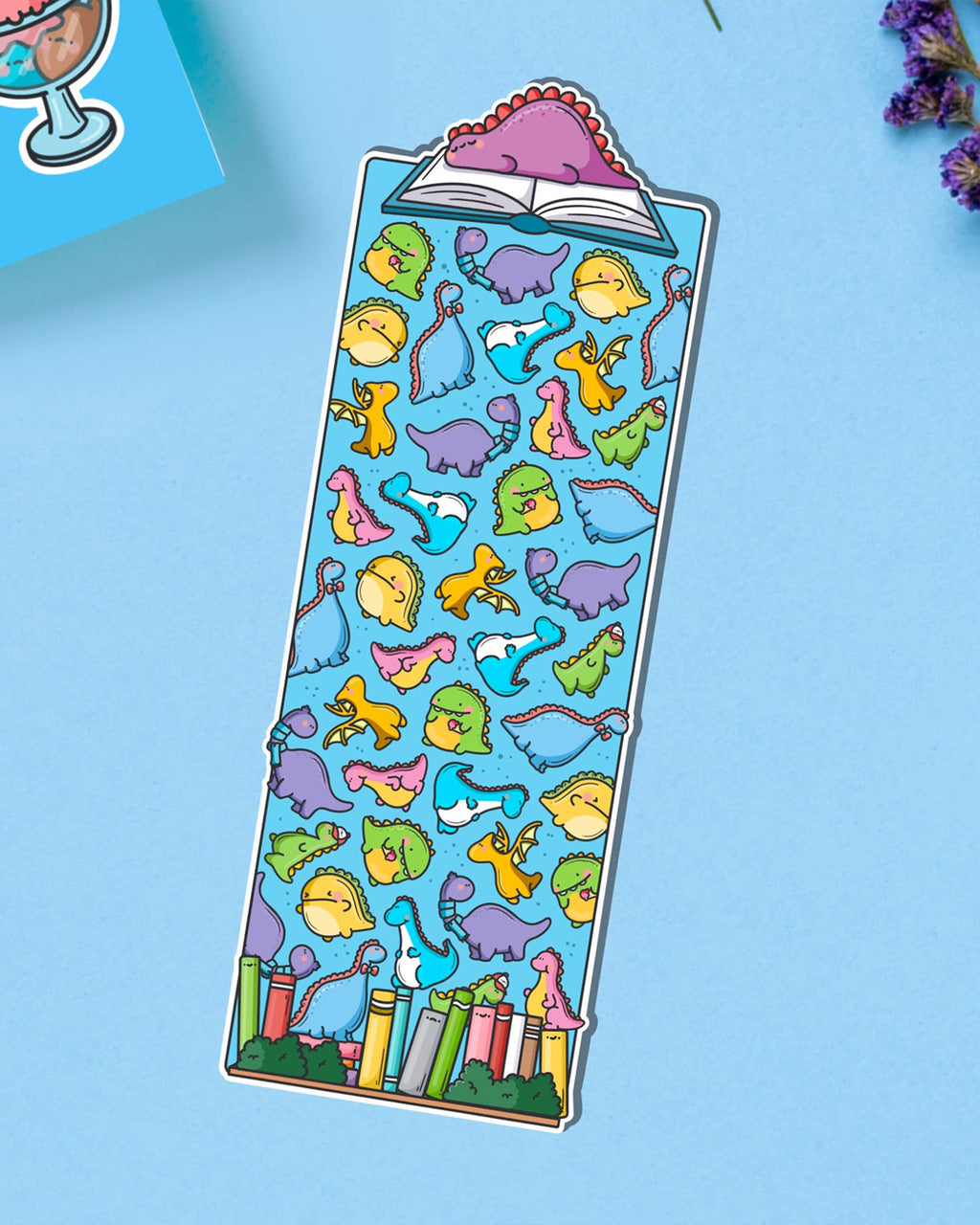 Fun dinosaur-themed bookmark designed by Toastedink, stocked at Rebel Romance, showing playful dinos in bright colours over a blue background. Photographed on a pastel blue surface with lavender nearby.