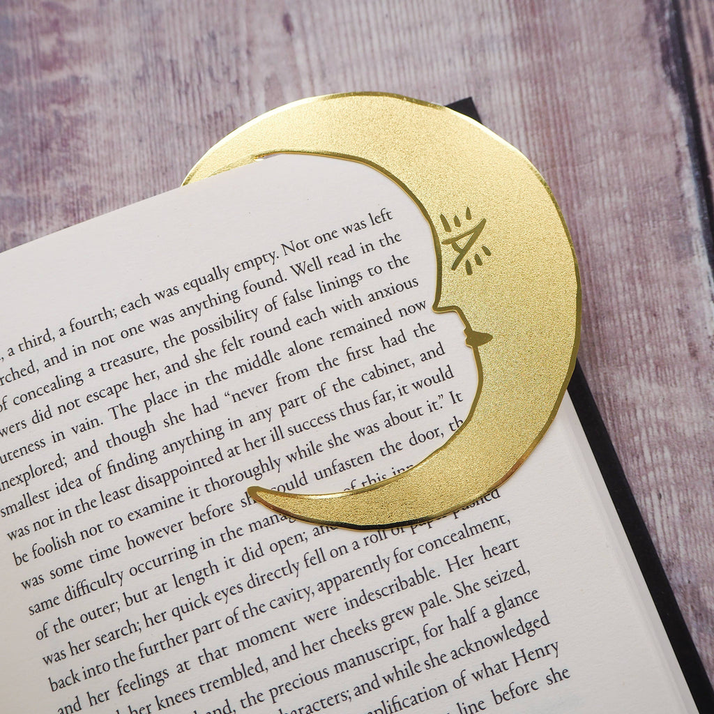 A crescent moon-shaped brass bookmark with “Midnight Readers Club” and celestial stars etched onto its surface, placed on an open book to mark a page. The bookmark adds an elegant and whimsical touch, ideal for book lovers who enjoy night-time reading.