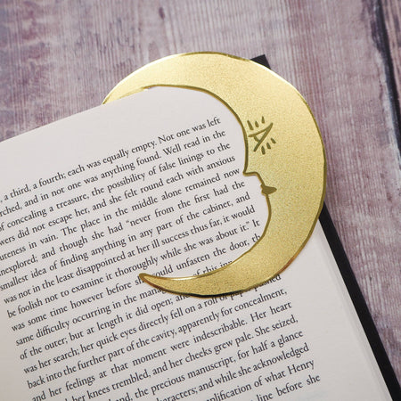 A crescent moon-shaped brass bookmark with “Midnight Readers Club” and celestial stars etched onto its surface, placed on an open book to mark a page. The bookmark adds an elegant and whimsical touch, ideal for book lovers who enjoy night-time reading.