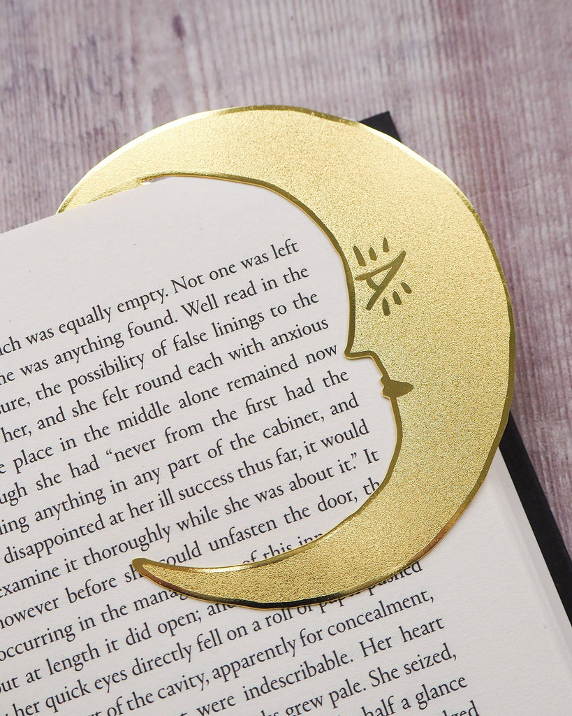 A crescent moon-shaped brass bookmark with “Midnight Readers Club” and celestial stars etched onto its surface, placed on an open book to mark a page. The bookmark adds an elegant and whimsical touch, ideal for book lovers who enjoy night-time reading.