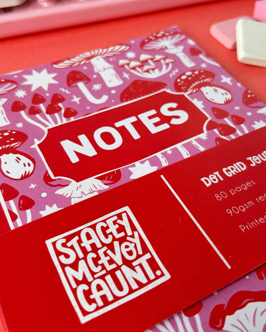 Close-up of the Stacey McEvoy Caunt pink and red mushroom notebook sold by Rebel Romance. The word "NOTES" is printed in bold white text on a red label, surrounded by a whimsical pattern of red and white toadstools on a pink background.