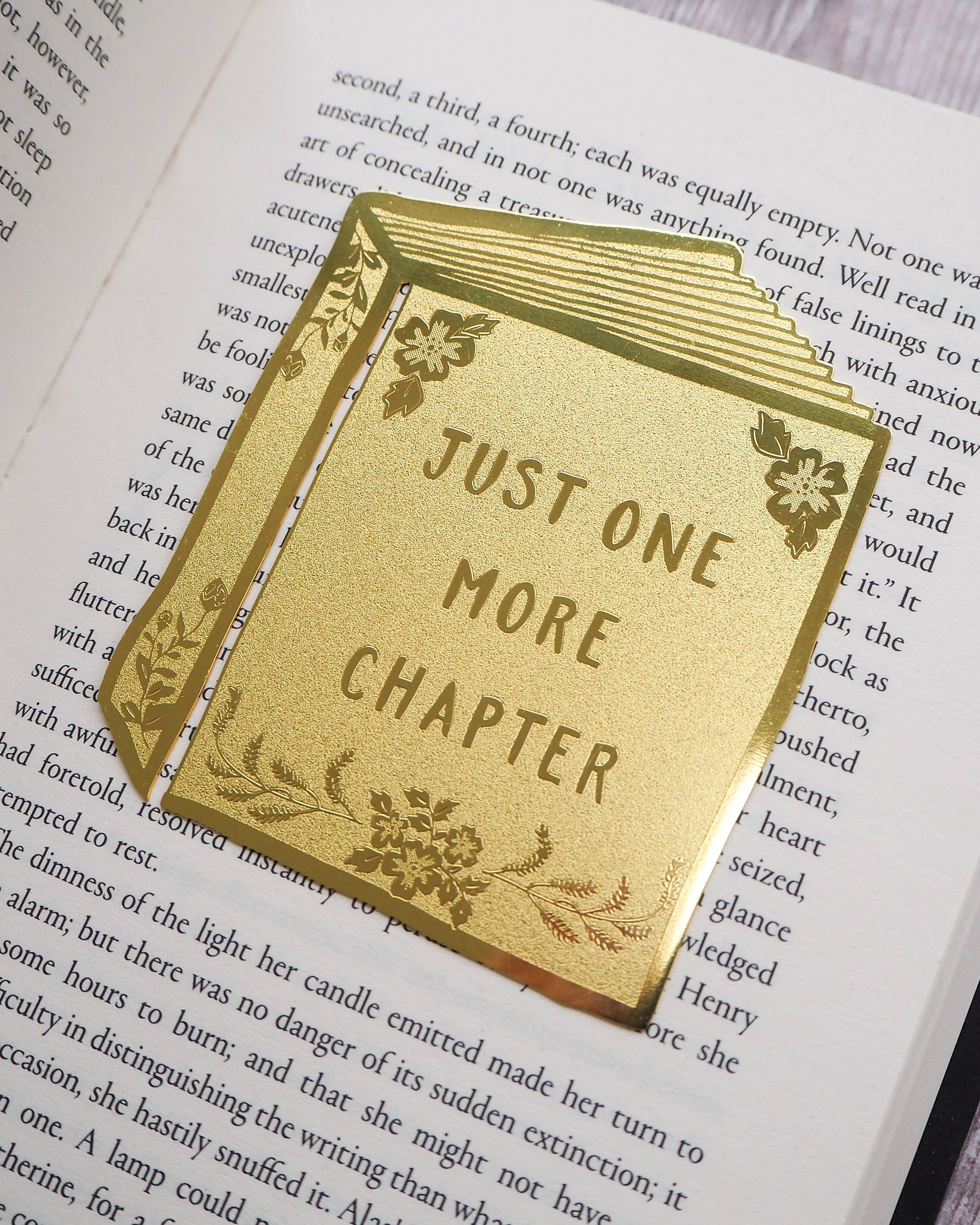 Close-up of a Literary Emporium brass bookmark reading “Just One More Chapter” with engraved floral accents. Photographed resting on an open book. Sold by Rebel Romance, a UK indie brand offering alternative gifts, homeware and slow ethical fashion.