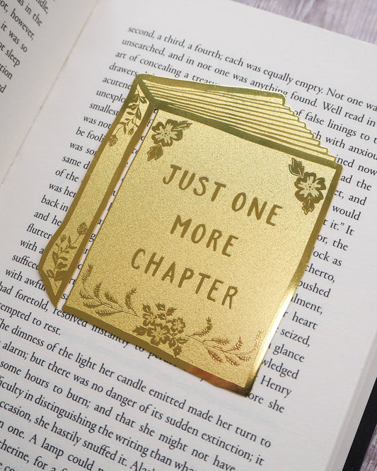 Close-up of a Literary Emporium brass bookmark reading “Just One More Chapter” with engraved floral accents. Photographed resting on an open book. Sold by Rebel Romance, a UK indie brand offering alternative gifts, homeware and slow ethical fashion.