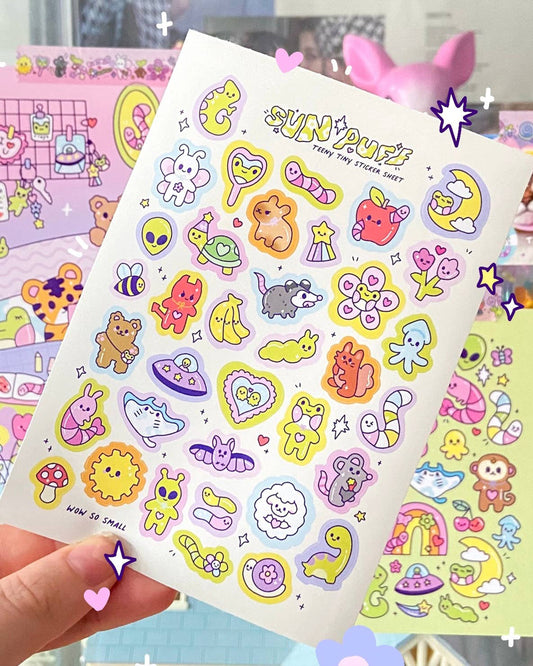 A6 sticker sheet by Sun Puff Studios, sold at Rebel Romance, featuring colourful teeny tiny vinyl stickers of cute characters like animals, UFOs, fruit, and stars. Each sticker is in a pastel palette of pink, yellow, purple and blue with kawaii faces, ideal for journaling or scrapbooking. Handheld for scale with other pastel sticker sheets visible in the background. Perfect for millennial stationery lovers who adore whimsical designs.