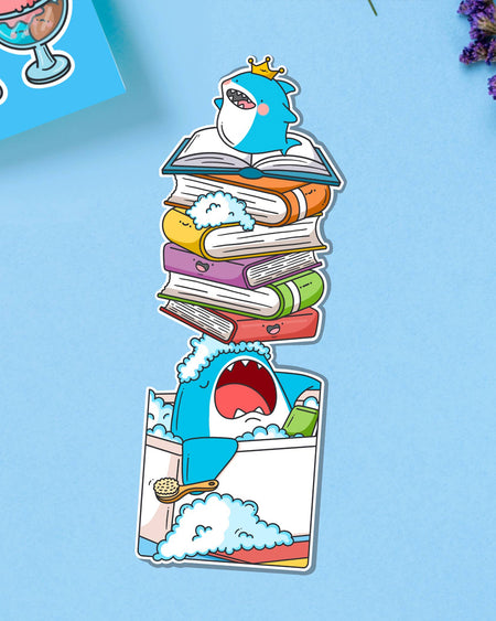 Adorable shark bookmark by Toastedink at Rebel Romance, with a cheeky blue shark in a bubbly tub and a rainbow stack of books on its nose. Pictured on a light blue background with purple flowers.