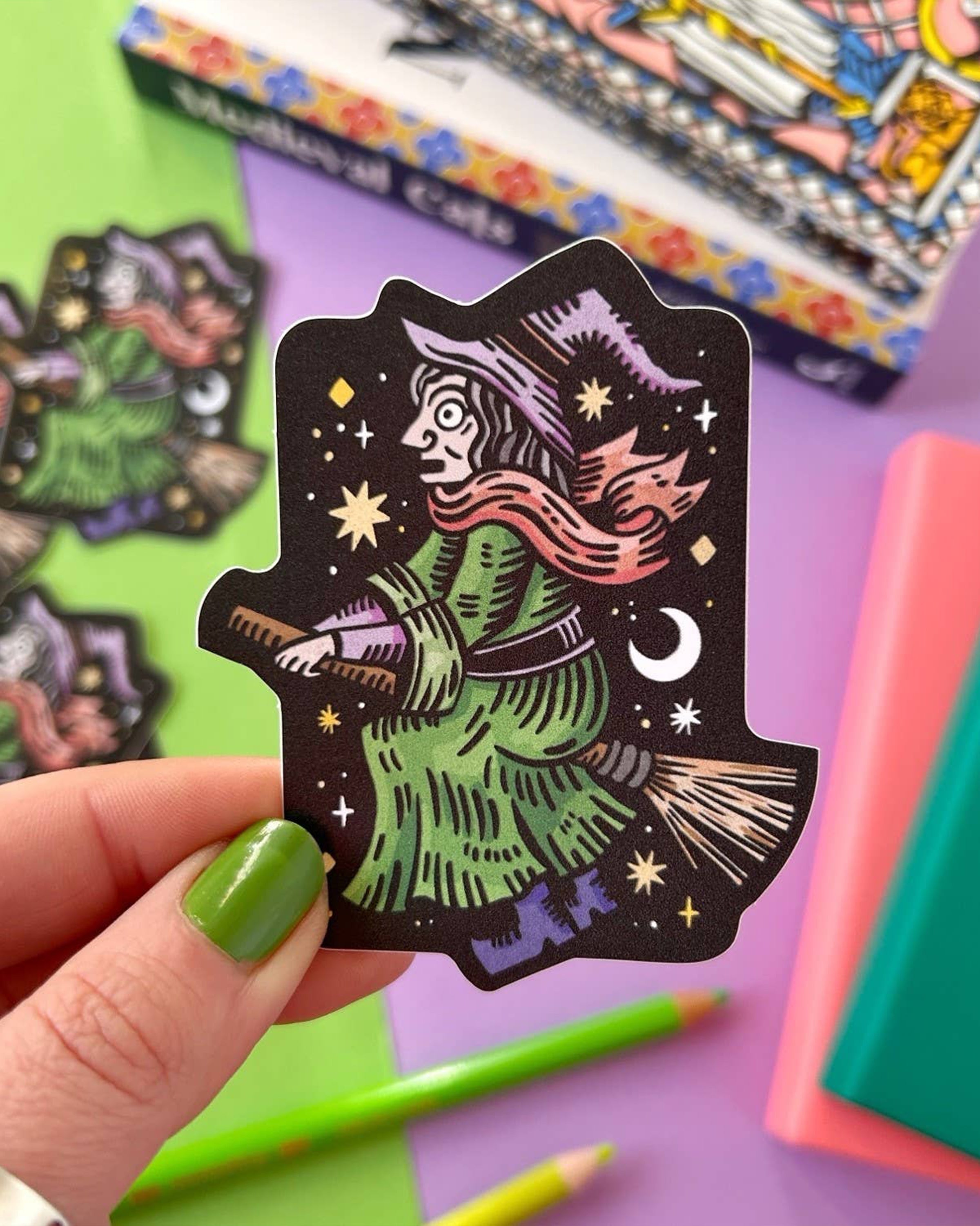 Close-up of a hand holding the witch on a broomstick vinyl sticker with a colourful background of pastel books, bright pencils, and a book titled 
