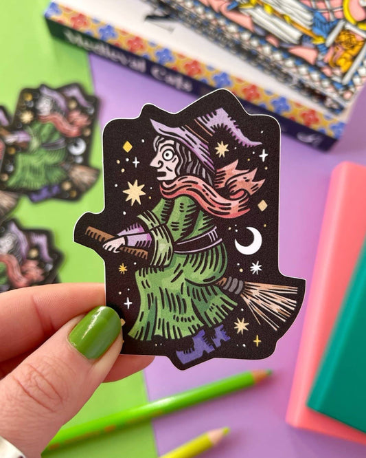 Close-up of a hand holding the witch on a broomstick vinyl sticker with a colourful background of pastel books, bright pencils, and a book titled "Medieval Cats". The sticker features a green-robed witch with a purple hat flying through a night sky. A magical, gothic accessory from Rebel Romance for alternative millennials.
