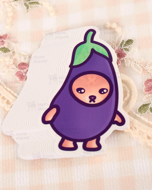 Stack of Rebel Romance Eggplant Cat vinyl stickers by Mona Mauve, featuring a sad cartoon cat dressed as a purple aubergine with a green leafy top. Part of the Snack Cats collection, this waterproof and durable sticker is ideal for decorating laptops, water bottles, or journals. Set on a pastel lace and floral fabric background. A quirky and kawaii design for millennials who love food-themed humour and alternative stationery.