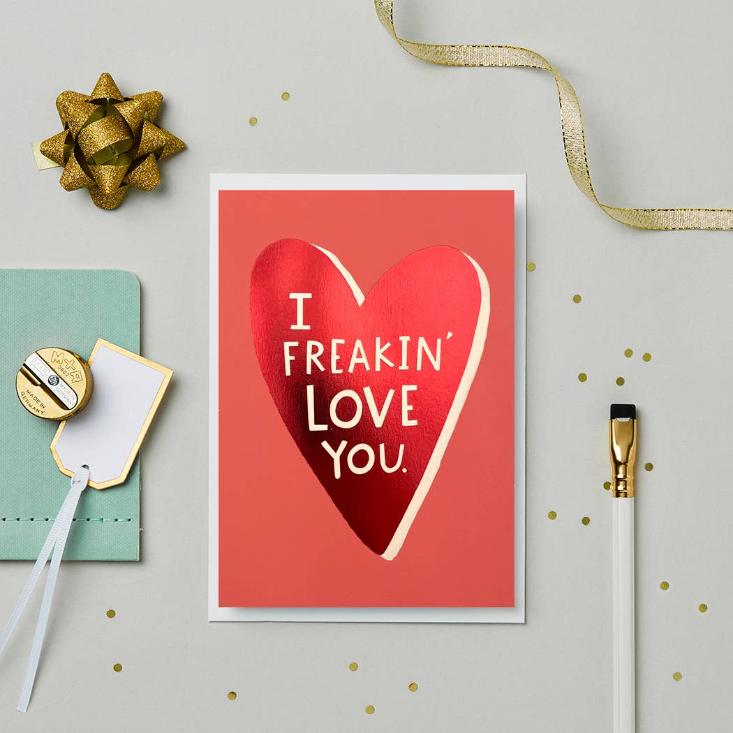 A close-up of the “I Freakin’ Love You” card featuring a shiny red foil heart and bold white text. The card is surrounded by gold and teal stationery accessories on a neutral background with gold confetti accents.