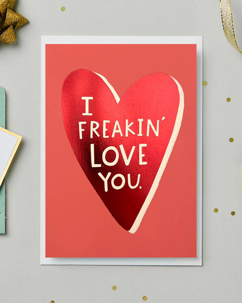 A close-up of the “I Freakin’ Love You” card featuring a shiny red foil heart and bold white text. The card is surrounded by gold and teal stationery accessories on a neutral background with gold confetti accents.