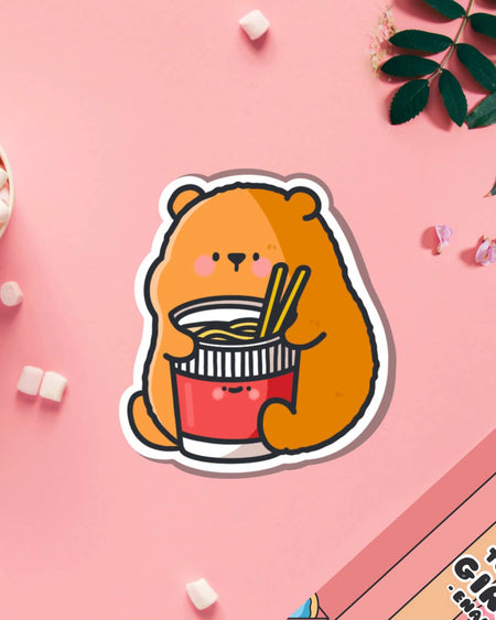 Cute vinyl sticker of a brown cartoon bear holding a cup of instant noodles with chopsticks, designed by Toastedink, placed on a pink background with marshmallows and petals.