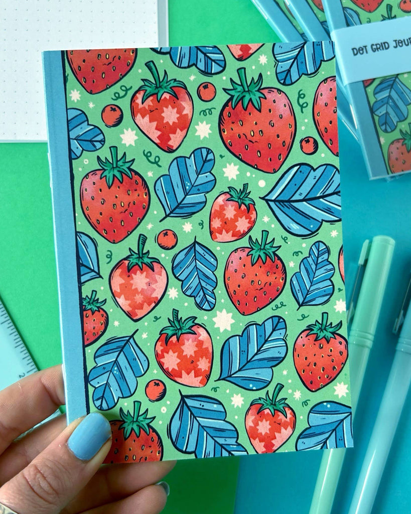 A hand holding the Rebel Romance Strawberry Pocket Notebook, showcasing its playful cover designed by Stacey McEvoy Caunt. Surrounded by pastel stationery, the bold red, green, and blue colours pop, ideal for creative journaling or gift giving.