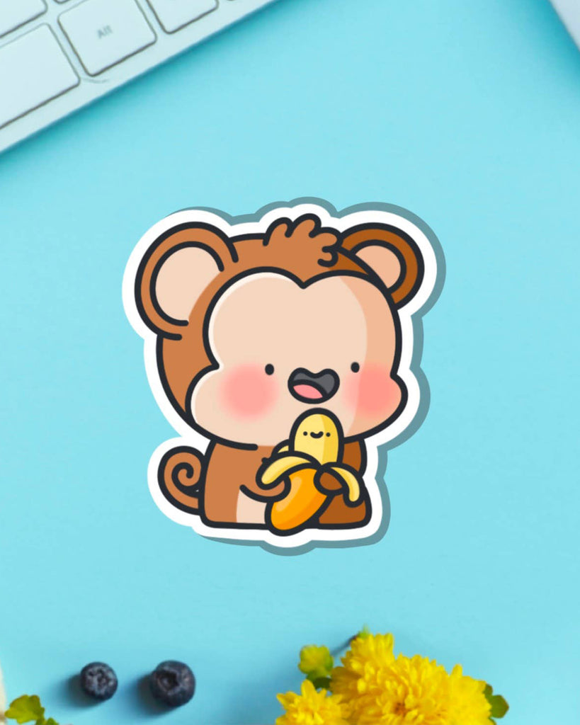 Whimsical monkey vinyl sticker with a happy banana on a bright turquoise desk background. A fun Toastedink design stocked by Rebel Romance, perfect for decorating laptops or notebooks.