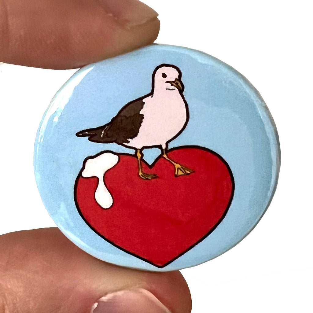 Close-up of the Rebel Romance "I Love Seagulls" button pin badge by Bite Your Granny, held between two fingers. The design features a cartoon seagull standing on a bright red heart with white seagull poop on it, set against a sky-blue background. A cheeky and fun seaside-inspired accessory from a UK small business celebrating alternative fashion and aesthetics.