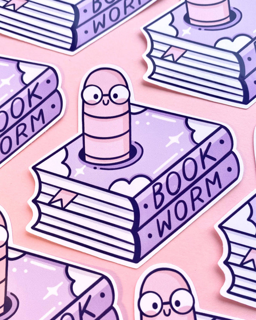 Flat lay of multiple Bookworm Glossy Stickers by Kelly Lou for Rebel Romance, featuring a happy pink worm with glasses inside a purple illustrated book labelled “BOOKWORM.” Displayed on a soft pink backdrop, these cute reading-themed vinyl stickers are ideal for bookworms, students, or anyone who loves fun stationery.