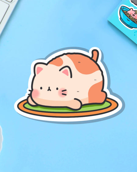 Toastedink's Resting Cat Vinyl Sticker, stocked by Rebel Romance. Features a cute loaf-shaped cat with peachy orange patches, relaxing on a rug. Displayed on a pale blue background next to a keyboard and postcard. Ideal for lovers of alternative, kawaii stationery and feline fun.
