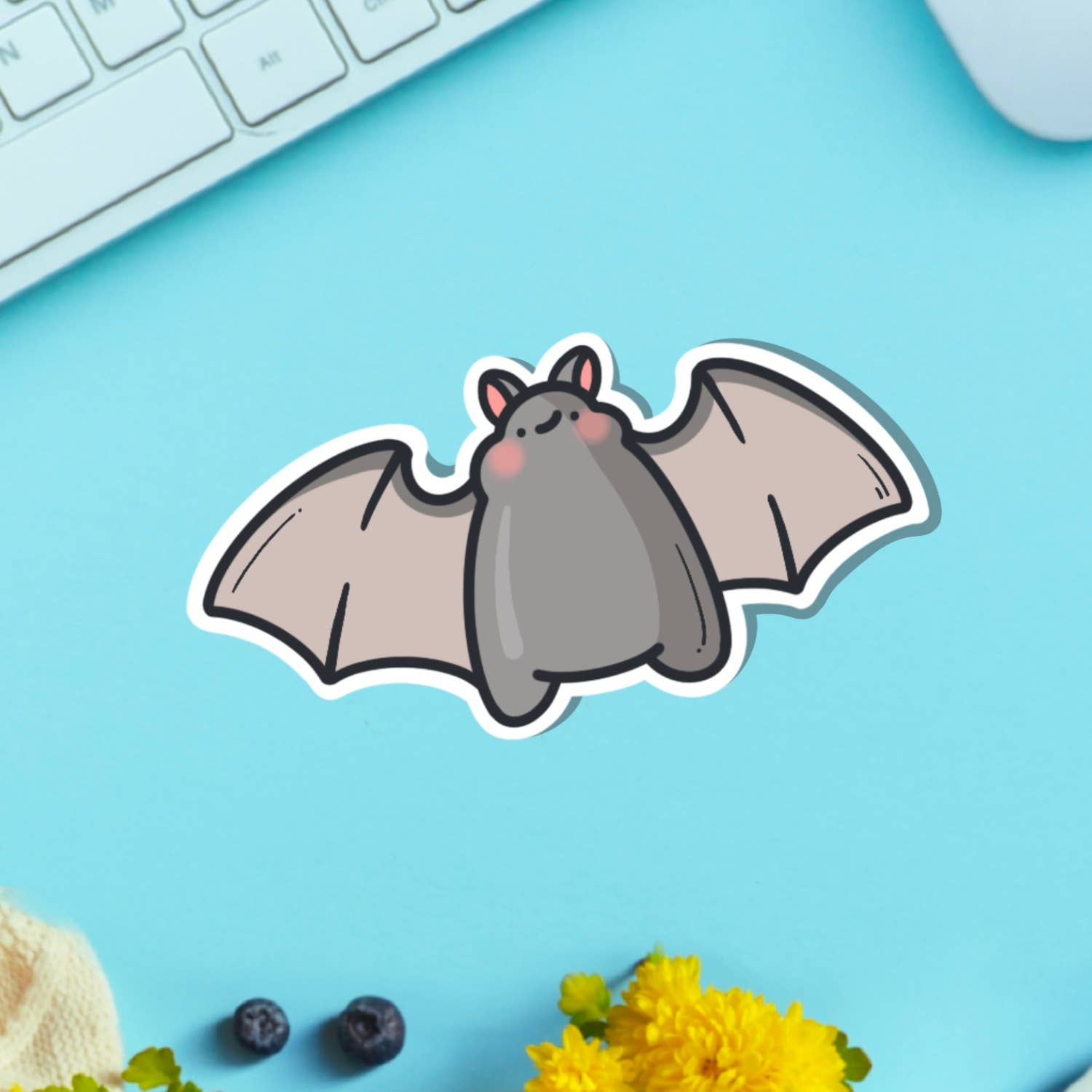 A cute vinyl sticker of a small grey cartoon bat with pink cheeks and a sweet expression, wings outstretched. Shown on a pink background with marshmallows and flowers. A waterproof, glossy sticker perfect for laptops, notebooks, or water bottles.