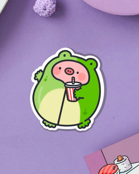 Vinyl sticker of a cute pig character wearing a green frog costume and sipping a red striped drink, photographed on a purple surface. Perfect for decorating laptops, bottles, or journals. Designed by Toastedink and stocked by Rebel Romance, a UK small biz celebrating quirky, alternative style.