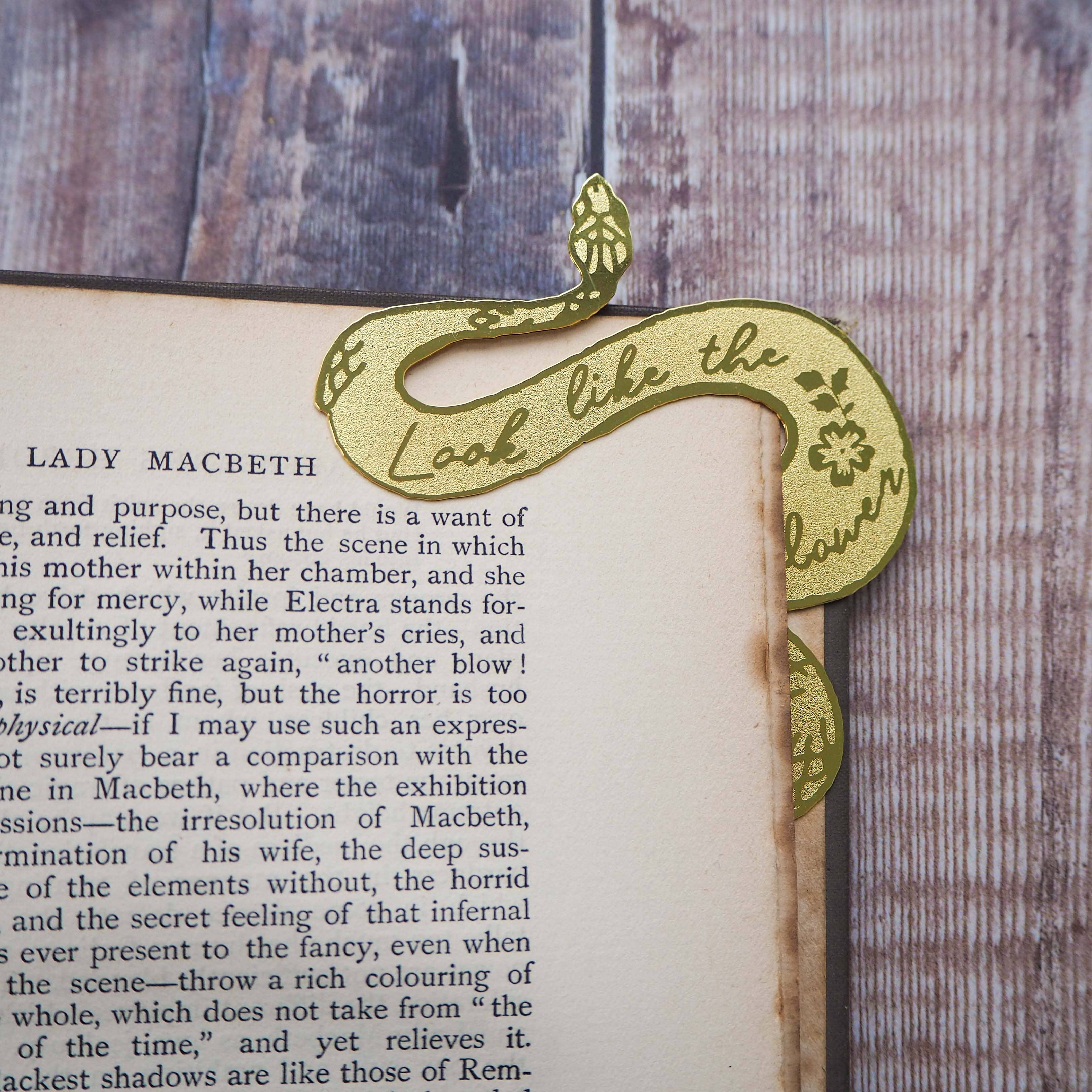 A brass bookmark shaped like a coiled serpent, delicately etched with the famous Lady Macbeth quote: “Look like the innocent flower, But be the serpent under ‘t.” The bookmark is placed inside an open book on a wooden surface, with the title “Lady Macbeth” visible on the page. The snake’s head is designed to peek out from the top of the book when closed, adding a dramatic and literary flair.