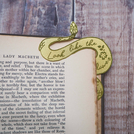 A brass bookmark shaped like a coiled serpent, delicately etched with the famous Lady Macbeth quote: “Look like the innocent flower, But be the serpent under ‘t.” The bookmark is placed inside an open book on a wooden surface, with the title “Lady Macbeth” visible on the page. The snake’s head is designed to peek out from the top of the book when closed, adding a dramatic and literary flair.