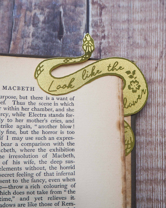 A brass bookmark shaped like a coiled serpent, delicately etched with the famous Lady Macbeth quote: “Look like the innocent flower, But be the serpent under ‘t.” The bookmark is placed inside an open book on a wooden surface, with the title “Lady Macbeth” visible on the page. The snake’s head is designed to peek out from the top of the book when closed, adding a dramatic and literary flair.