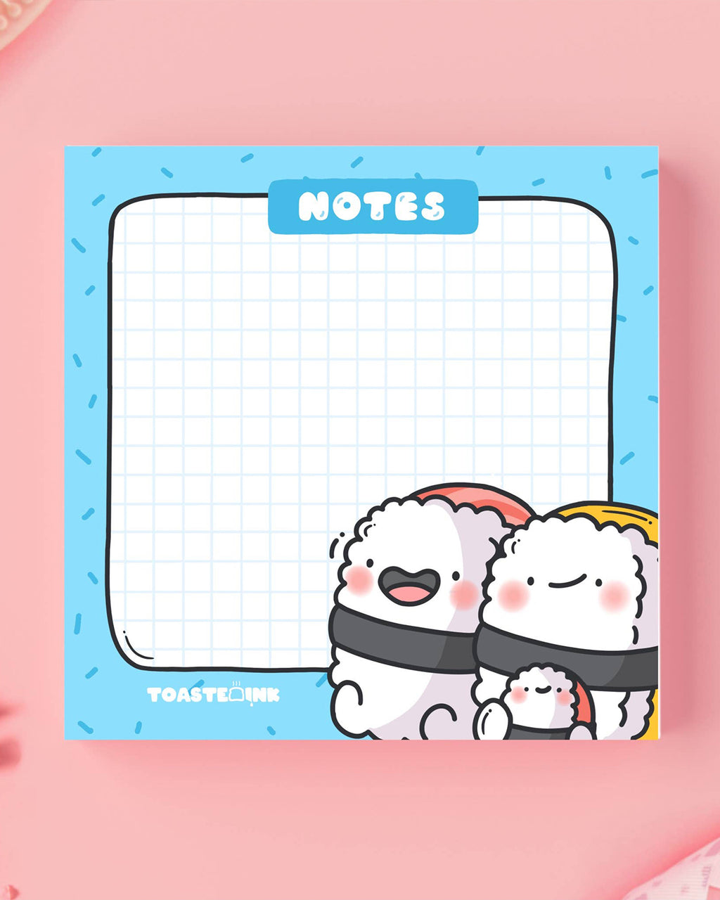 Toastedink Sushi Sticky Notes styled on a pink surface, featuring happy sushi characters at the bottom and a grid for writing. Stocked by Rebel Romance, a UK brand for quirky stationery.