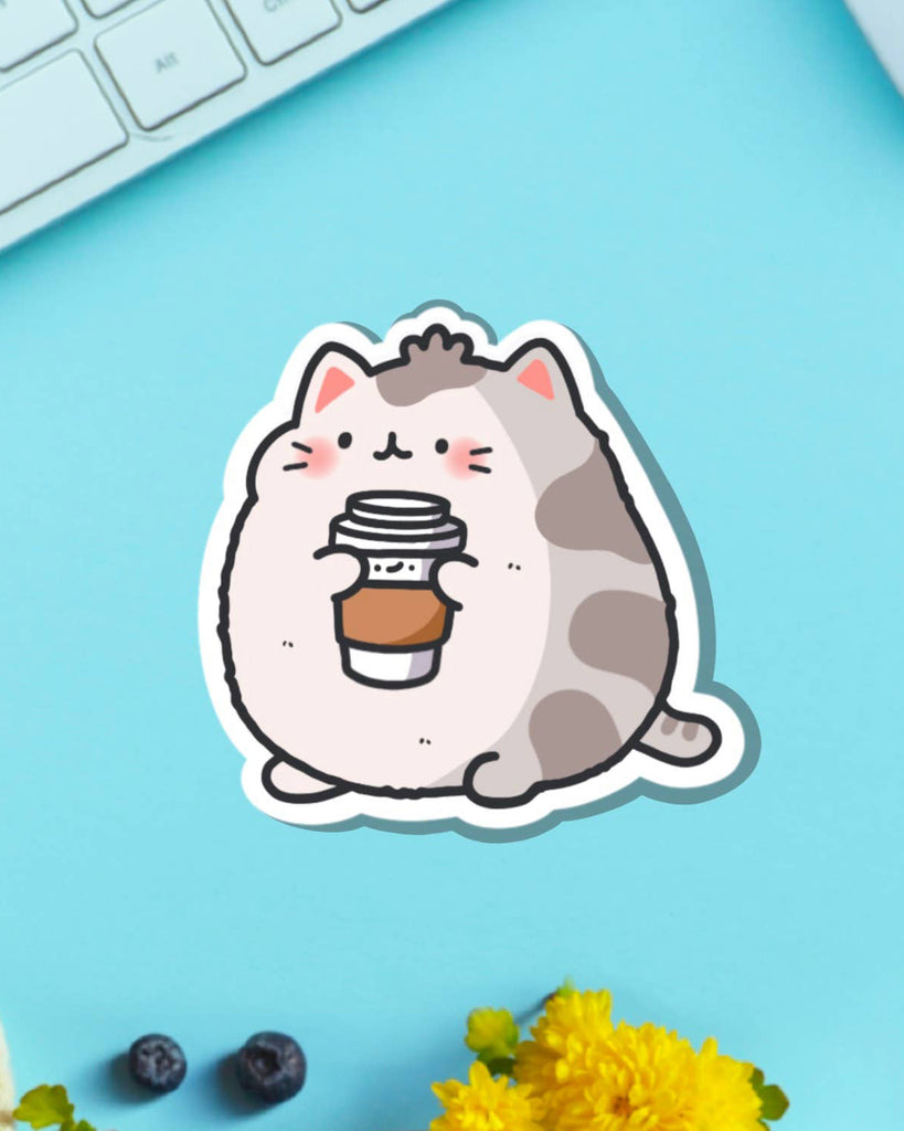 Toastedink Coffee Cat Sticker placed on a blue background with a keyboard and flowers. A cute grey and white cat clutches a cup of coffee with a sleepy face. Ideal for decorating notebooks, planners, or as a gift for coffee and cat lovers.